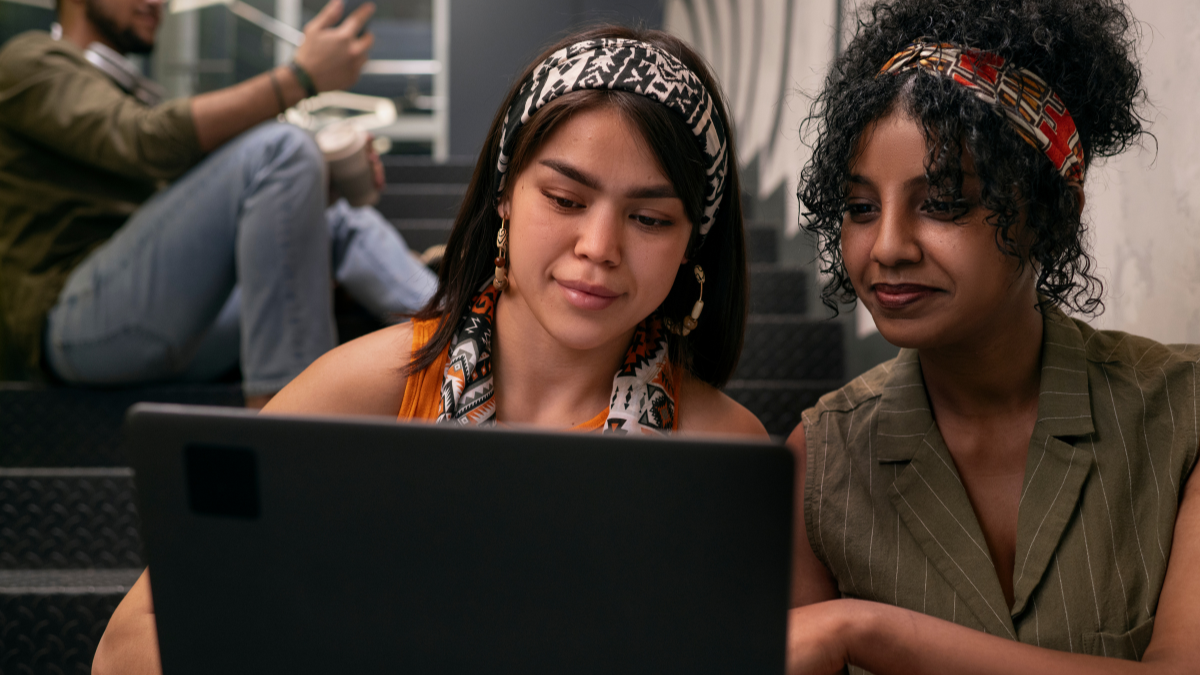 Duas mulheres em um ambiente de escritório, analisando informações no notebook. Elas usam faixas estampadas no cabelo e parecem concentradas durante uma conversa tranquila.