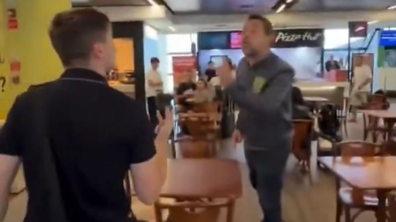 Lucas Pavanato e Guilherme Boulos conversam em um ambiente interno, como restaurante ou pizzaria, enquanto uma pessoa em primeiro plano segura o celular e o outro observa ao fundo. Mesas e cadeiras visíveis.