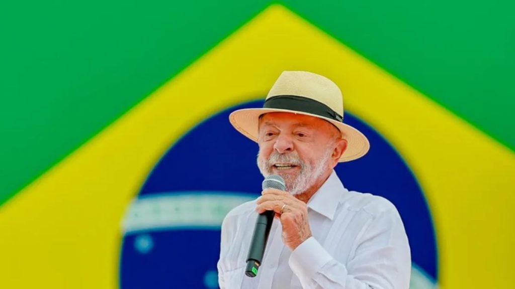 Presidente Lula fala ao microfone diante de uma bandeira do Brasil ao fundo.