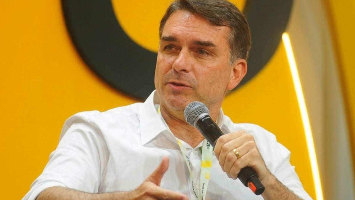 Flávio Bolsonaro segura um microfone e gesticula durante participação em evento.