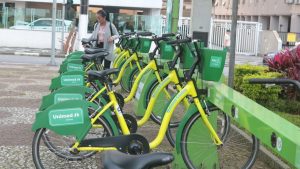 Bicicletas compartilhadas verdes e amarelas estacionadas ao lado de um prédio, com uma pessoa ao fundo usando celular, com lixeiras Unimed ao longo da área