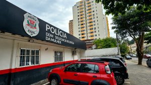 Prédio da Polícia Civil com fachada “DDM da Mulher” e viatura estacionada em frente, em área urbana com prédios ao fundo.