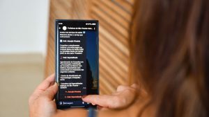 Pessoa usando um celular com tela de aplicativo de informações sobre serviços e atendimento em saúde, com opções e mensagens de orientação exibidas no visor.