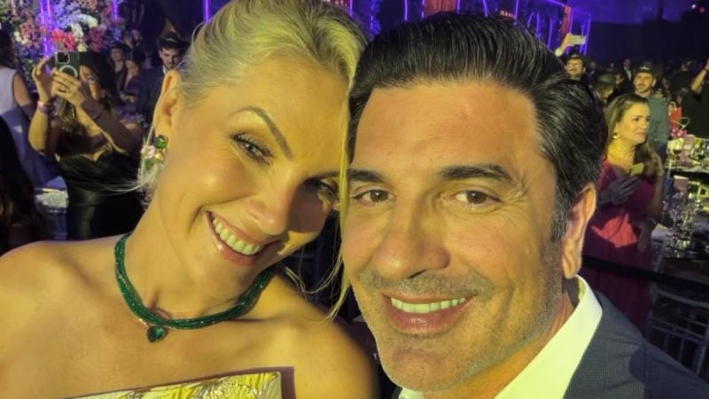Foto de casal em evento à noite: Ana Hickmann sorri ao lado de Edu Guedes que está de terno, com iluminação roxa ao fundo e público ao redor.