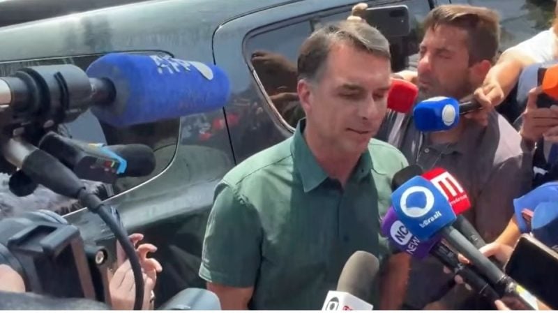 Flávio Bolsonaro de camisa verde é entrevistado por jornalistas com vários microfones e repórteres do lado de fora, em frente a um carro, em cobertura ao vivo.