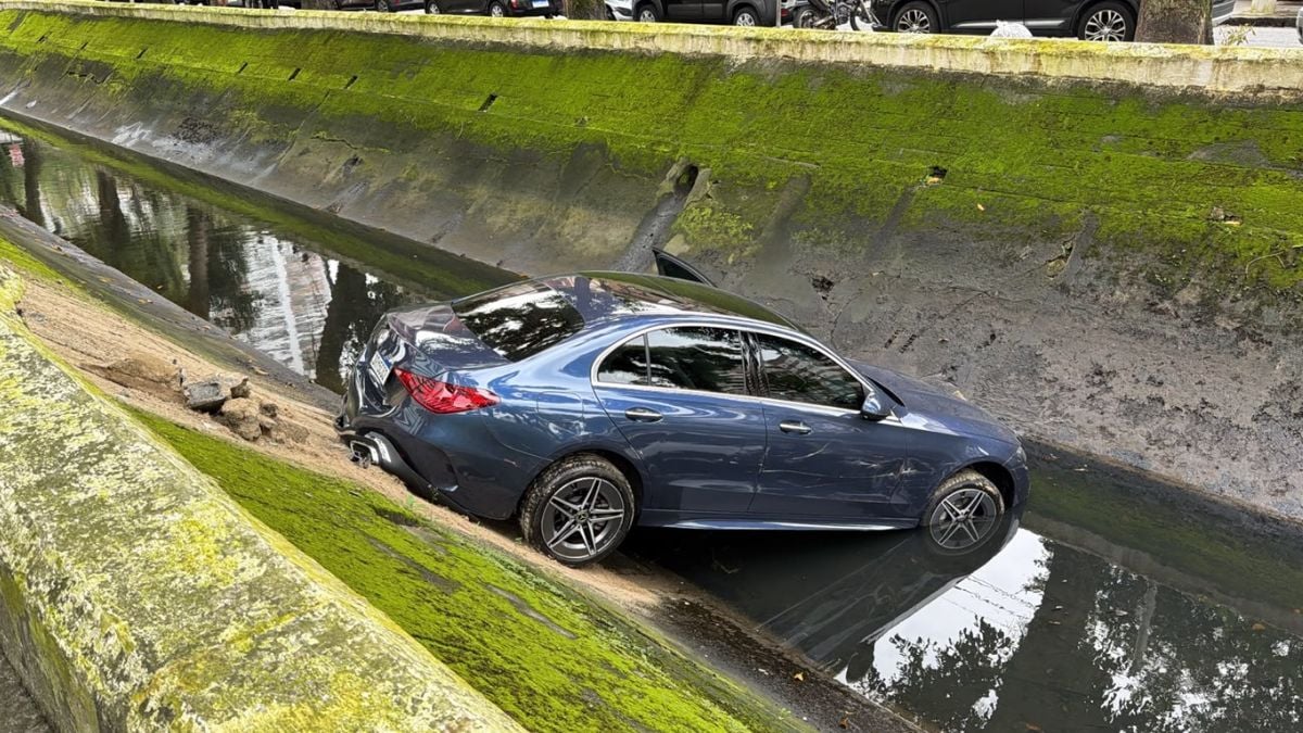 Sedã azul em manobra em uma pista de concreto com água ao lado, exibindo rodas com aro esportivo e reflexo no chão molhado durante um evento automotivo.