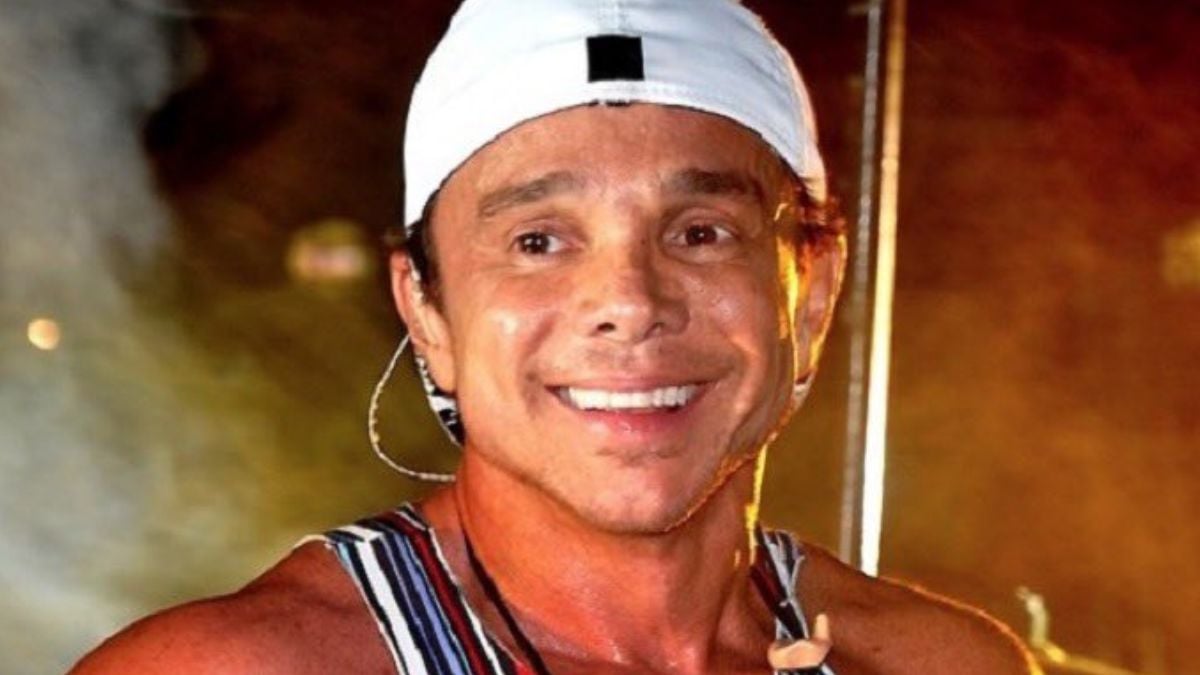 Netinho sorrindo em uma cena noturna, com boné branco e jaqueta listrada, ao lado de um ambiente externo desfocado.