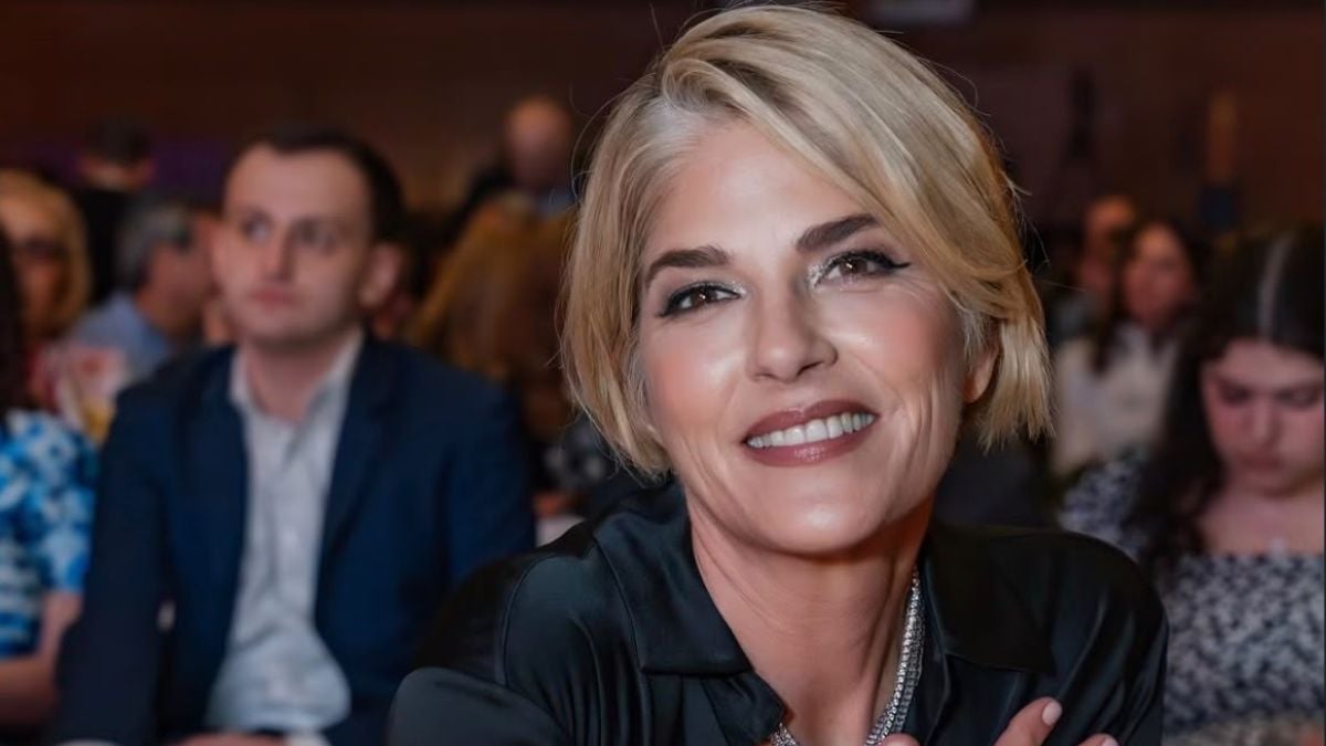 Foto de Selma Blair loira de cabelo curto, usando maquiagem e blusa escura, sorrindo em um evento com pessoas ao fundo em ambiente interno.