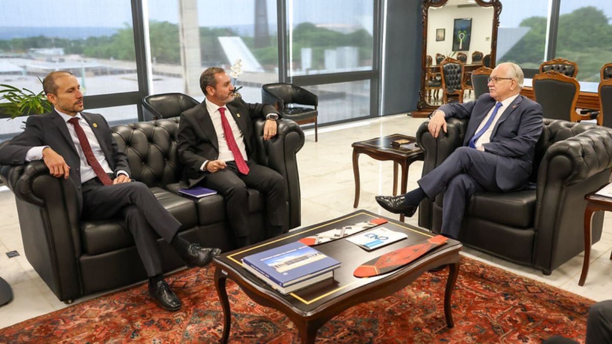 Reunião em sala executiva com três homens em sofás de couro, um deles de óculos, conversando durante encontro oficial; mesa de centro com pastas e documentos.