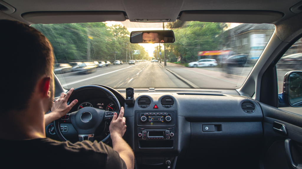 Vista em perspectiva de dentro de um carro em movimento, com o motorista ao volante e o painel, enquanto a estrada urbana segue à frente ao entardecer.