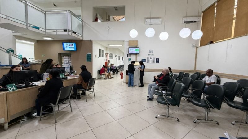Sala de atendimento em um Mutirão da Saúde em Bragança Paulista, com pessoas sentadas e em pé, e estações de triagem ao fundo.