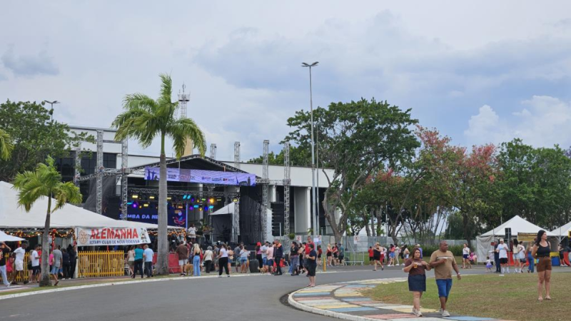 Área externa de evento com palco montado, torres de iluminação, tendas e pessoas circulando em frente a um complexo esportivo em dia de céu parcialmente nublado.