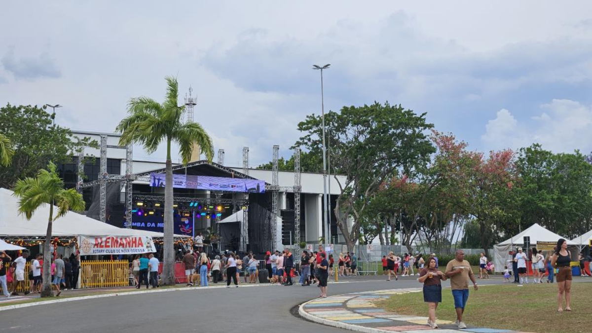 Área externa de evento com palco montado, torres de iluminação, tendas e pessoas circulando em frente a um complexo esportivo em dia de céu parcialmente nublado.