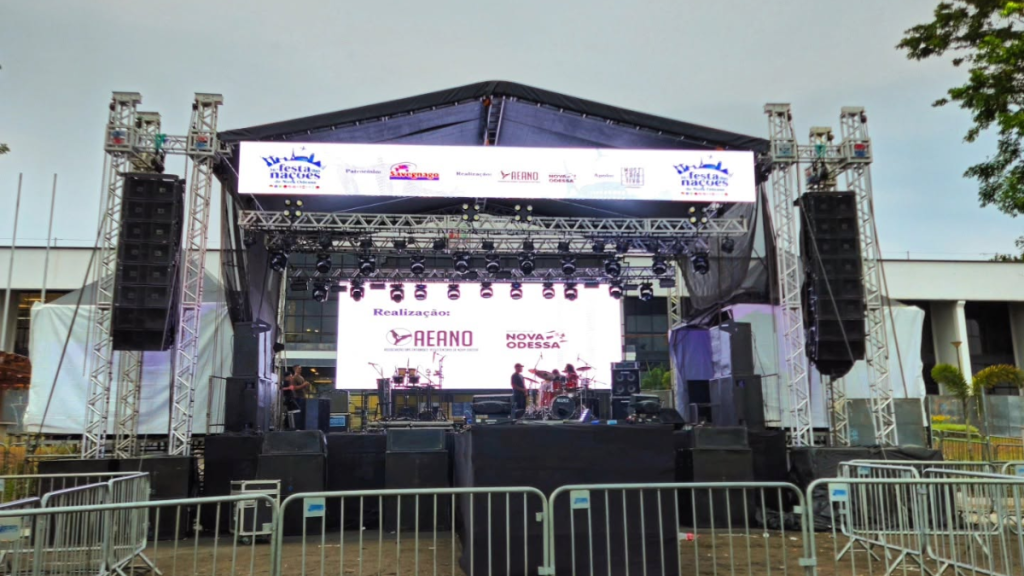 Vista de um evento ao ar livre com palco de shows, iluminação e tendas, com pessoas circulando em área externa e árvores ao fundo.
