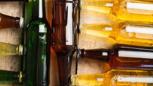 Garrafas de vidro com cervejas e bebidas alcoólicas em diferentes cores, com rótulos fechados e reflexos de luz, ambiente de bar ou adega, close-up