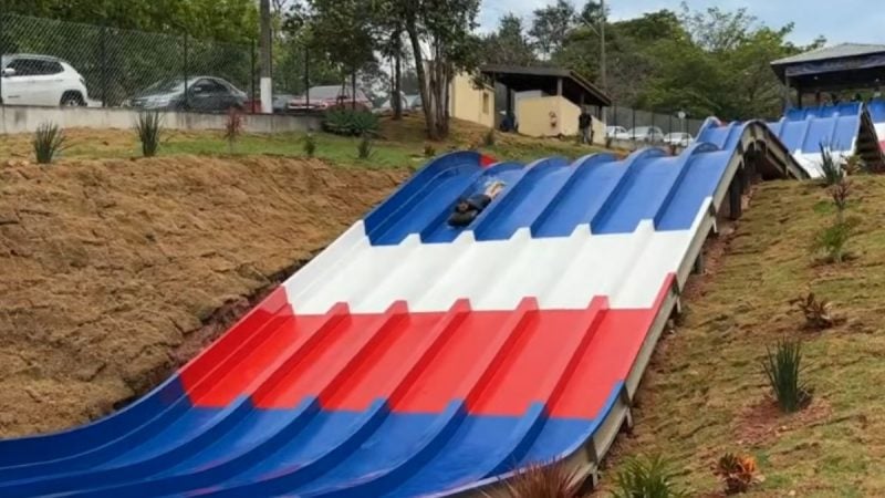 Parque aquático em Águas de Lindóia inaugura nova atração com tobogã duplo azul e vermelho em meio a área externa, céu nublado e vegetação ao redor.