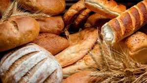 Pães artesanais e rústicos assados, incluindo pão redondo fatiado e rolinho com casca dourada, com trigo ao fundo, prontos para servir.