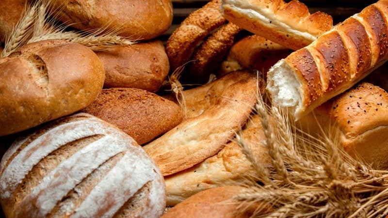 Pães artesanais e rústicos assados, incluindo pão redondo fatiado e rolinho com casca dourada, com trigo ao fundo, prontos para servir.