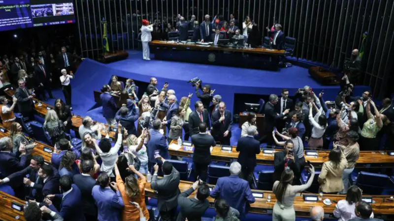 Câmeras e fotógrafos em pé no plenário do Congresso Nacional, com parlamentares na área de votação e equipe de imprensa registrando a sessão no Senado Federal