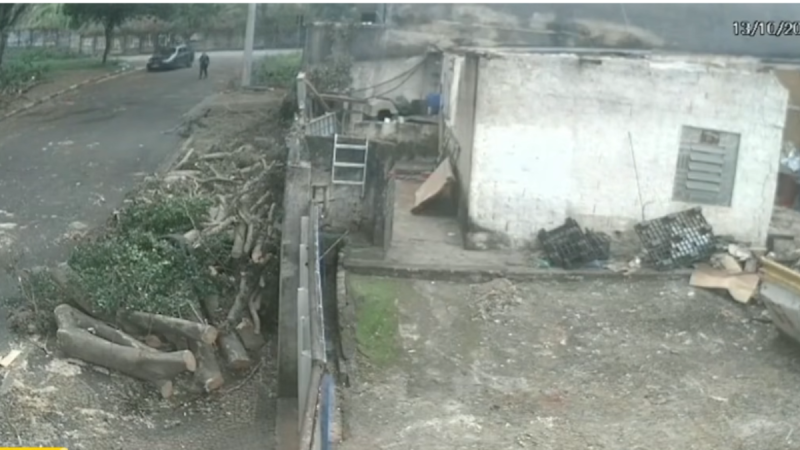 Imagem de videomonitoramento de um terreno em área rural, com pilhas de madeira cortada, cerca e um galpão branco parcialmente danificado, com sujeira e entulho no chão.