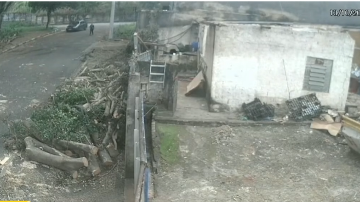 Imagem de videomonitoramento de um terreno em área rural, com pilhas de madeira cortada, cerca e um galpão branco parcialmente danificado, com sujeira e entulho no chão.