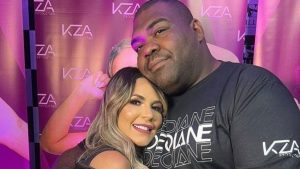 Nego Jader e Deolane posando em evento com fundo de estande roxo e logo KZA, com homem usando camiseta preta e mulher sorrindo ao lado, em clima de entrevista ou foto promocional