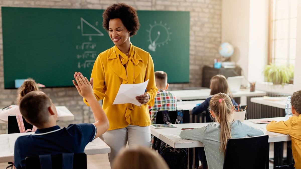 Professora sorri para a turma enquanto apresenta uma folha de atividades em sala de aula, com quadro verde ao fundo e estudantes sentados em carteiras, em uma cena educativa.