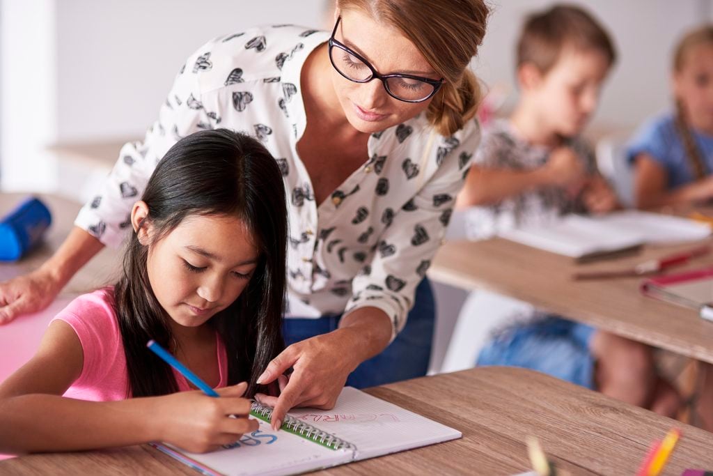 Professora ajuda criança a desenhar e colorir em uma sala de aula, enquanto outras pessoas trabalham ao fundo, em atividade pedagógica com caderno e lápis.