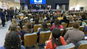 Público sentado em auditório durante o Feirão de Empregos de Hrtolândia, com palestrantes ao fundo em palco e cartazes informativos ao redor.