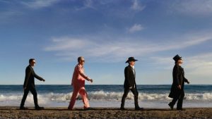 Quatro integrantes do U2 caminham pela praia à beira-mar ao pôr do sol, com o céu azul e nuvens, em imagem promocional de shows no Brasil.