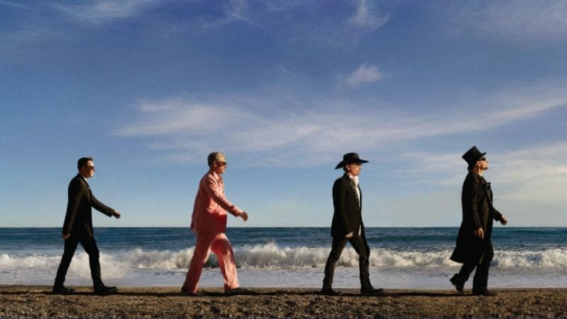 Quatro integrantes do U2 caminham pela praia à beira-mar ao pôr do sol, com o céu azul e nuvens, em imagem promocional de shows no Brasil.