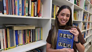 A professora de matemática Luana Rodrigues de Aguiar sorri e segura o livro “História da Matemática” em frente a estantes com livros.