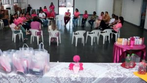 Encontro em ambiente interno com pessoas sentadas em cadeiras durante evento temático rosa, mesa decorada com lembrancinhas em sacolas e artigos em tons de rosa e branco.