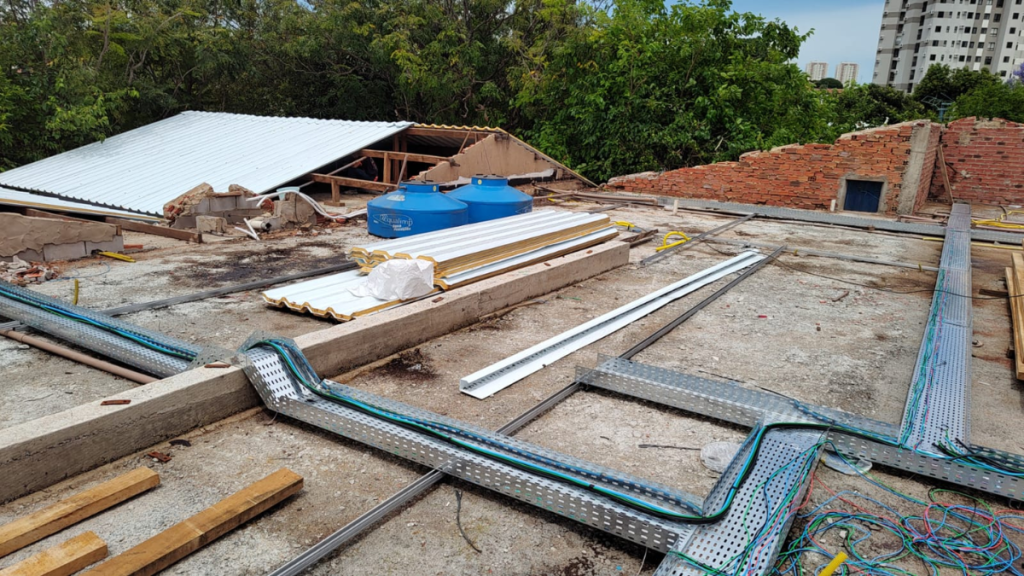 Vista de uma obra em andamento com telhado em construção, vigas e estruturas metálicas, canalizações e materiais sobre a laje, com vegetação ao fundo.