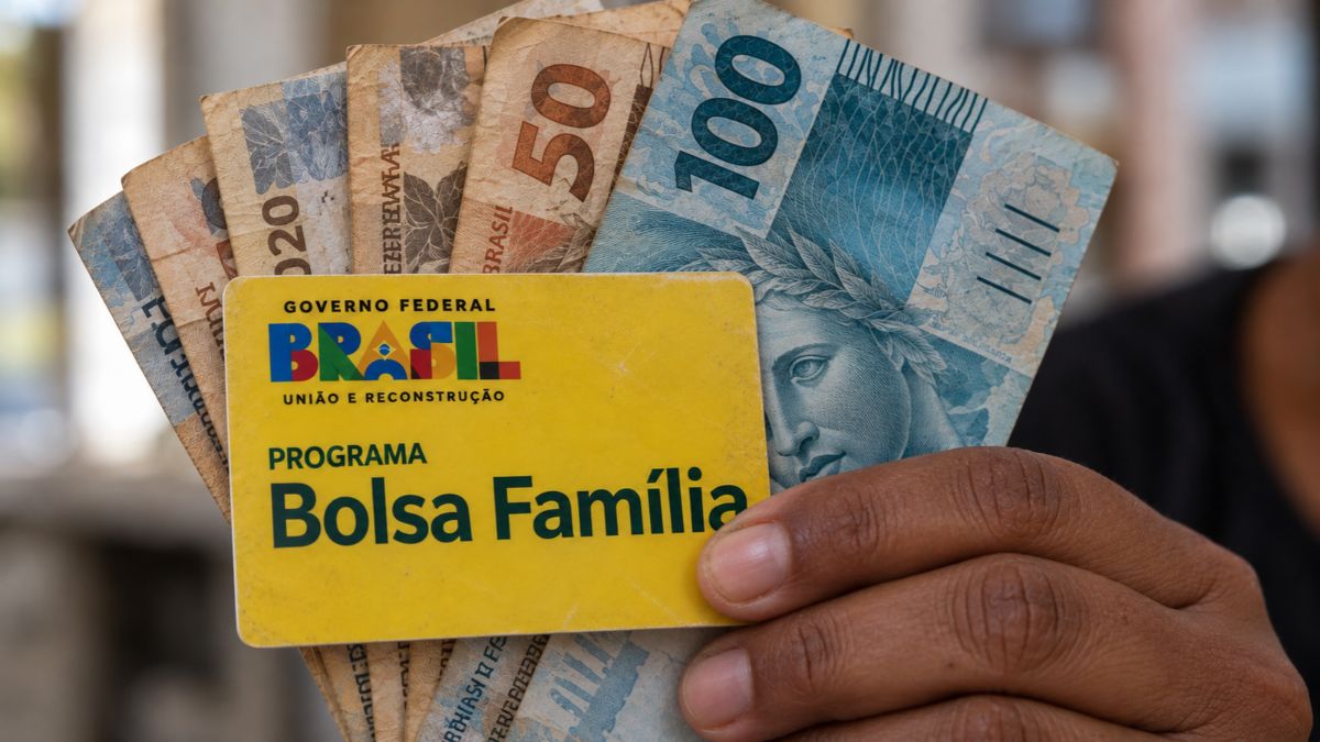 Reajuste do Bolsa Família: mão segura cartões do Programa Bolsa Família e notas de reais ao fundo, em imagem que simula informações sobre benefícios sociais.