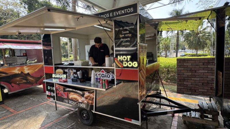Carrinho de hot dog e hambúrguer em evento ao ar livre, com atendente preparando lanches sob uma tenda, placa “HOT DOG” e decoração colorida do food truck.