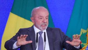 Lula em discurso, ao lado de bandeiras do Brasil e de outros símbolos, em um palco com microfones e expressão gesticulando com as mãos
