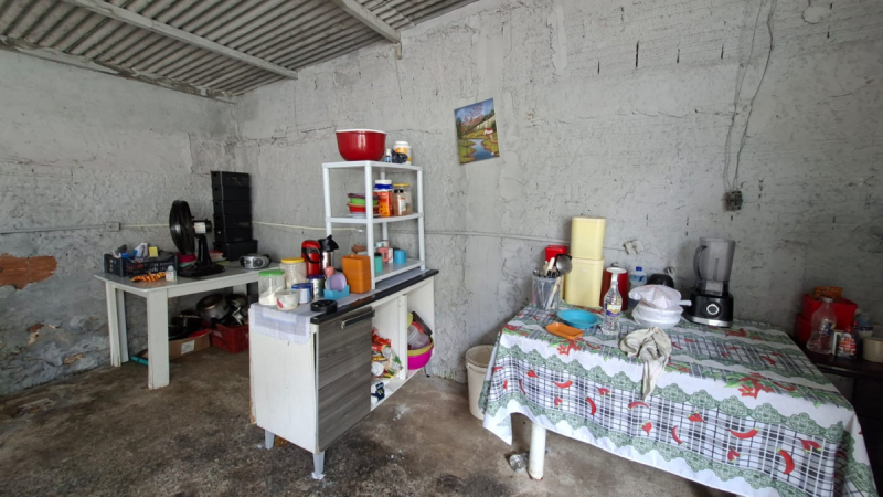 Cômodo com parede de cimento, mesa com toalha estampada e armário branco com prateleiras cheias de utensílios e produtos, em ambiente interno simples e organizado.