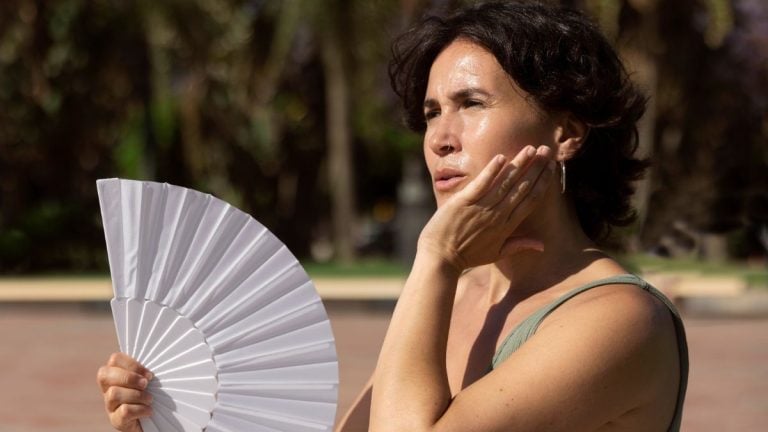 Mulher ao ar livre segura um leque branco e toca o rosto, em imagem que aborda Menopausa antes dos 45 anos: sinais e por que isso acontece.