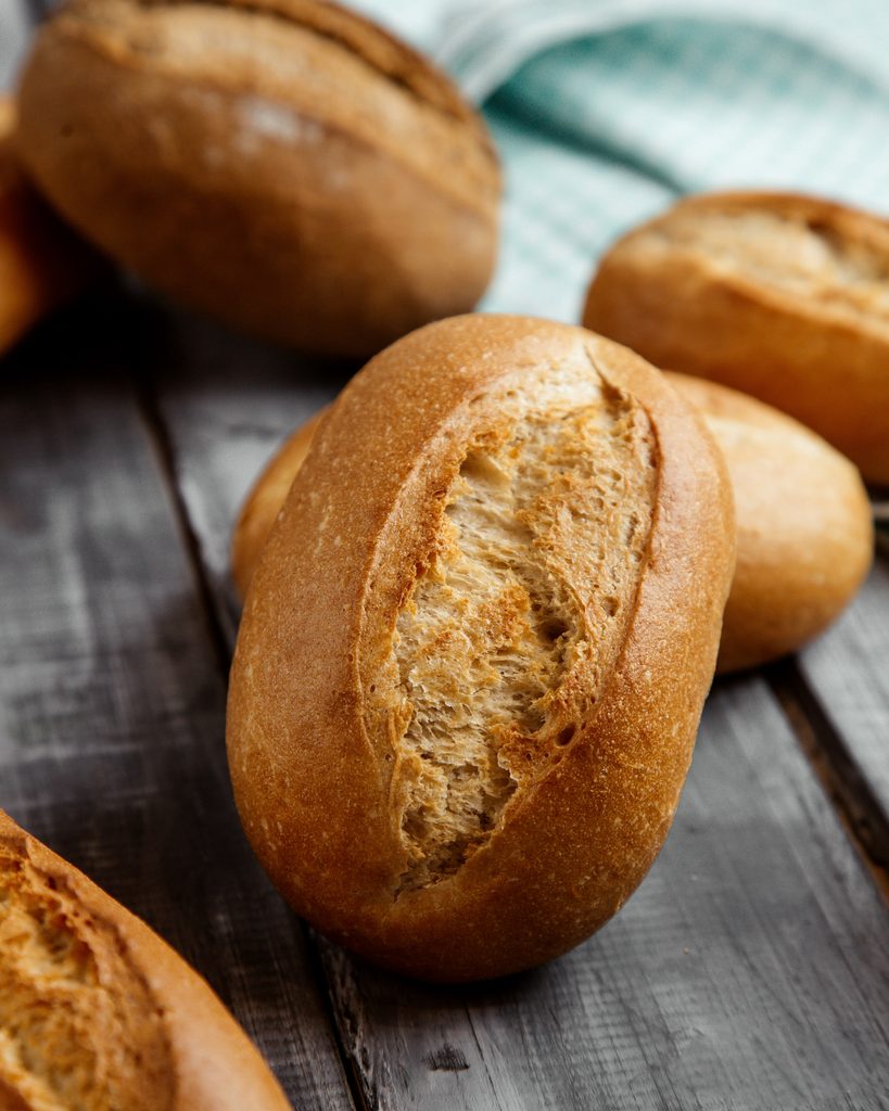 Pães caseiros crocantes e dourados, com miolo aerado e parte cortada, sobre uma tábua de madeira, com fundo desfocado em tecido claro ao lado.