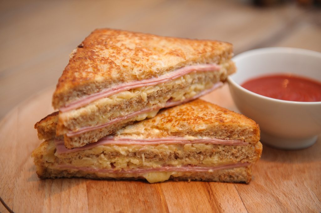 Misto quente na chapa, com fatias douradas e queijo derretido no meio, servido em tábua de madeira com molho em um potinho ao lado.