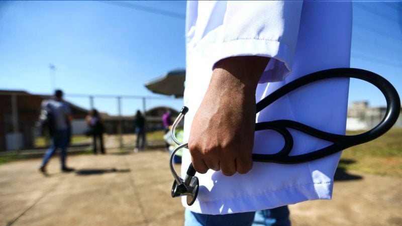 Médico em ambiente externo, segurando um estetoscópio com as mãos, usando jaleco branco e atendendo em área aberta com pessoas ao fundo.