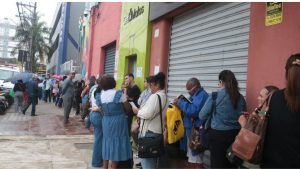 Pessoas em fila na calçada em frente a uma loja com porta verde e fachada rosa, com trabalhador segurando um documento e moradores aguardando atendimento na rua. Concurso público em Santos