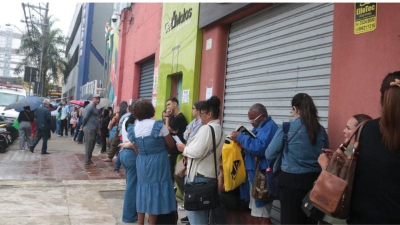 Pessoas em fila na calçada em frente a uma loja com porta verde e fachada rosa, com trabalhador segurando um documento e moradores aguardando atendimento na rua. Concurso público em Santos