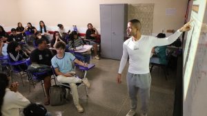 Professor em sala de aula com estudantes sentados em cadeiras, enquanto um aluno apresenta um cartaz ou material visual na frente da turma, em ambiente escolar.