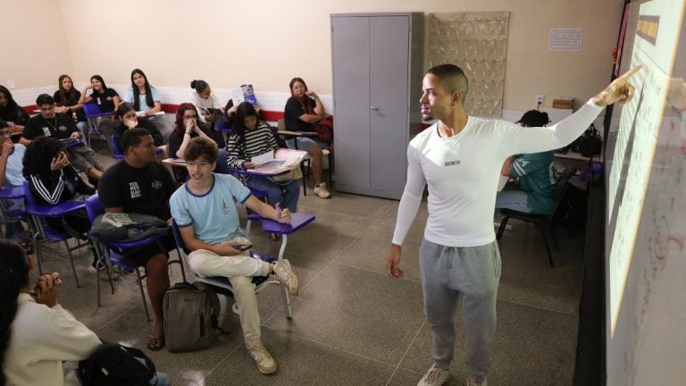 Professor em sala de aula com estudantes sentados em cadeiras, enquanto um aluno apresenta um cartaz ou material visual na frente da turma, em ambiente escolar.