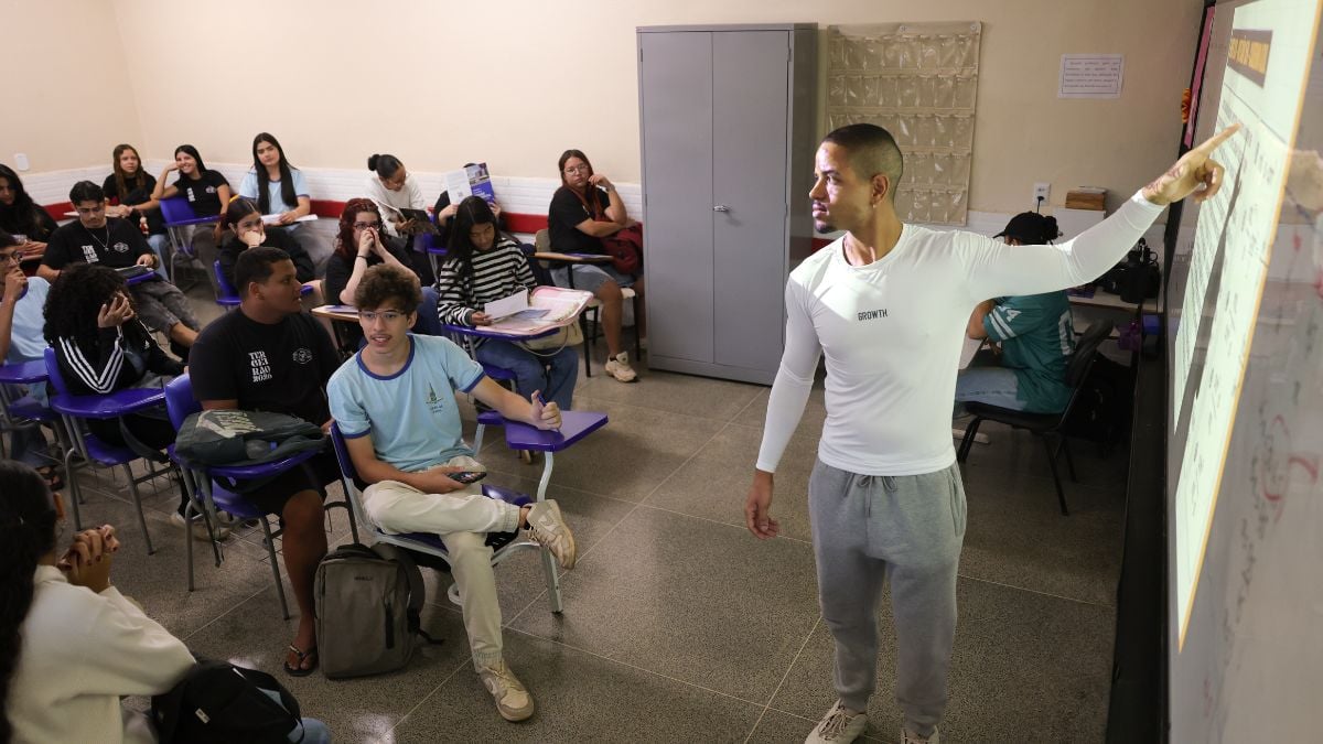Professor em sala de aula com estudantes sentados em cadeiras, enquanto um aluno apresenta um cartaz ou material visual na frente da turma, em ambiente escolar.