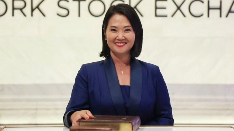 Foto de Keiko Fujimori em evento, usando blazer azul e expressão sorridente, posicionada atrás de um púlpito com painel ao fundo com a palavra “EXCHA”.