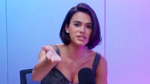 Bruna Marquezina em entrevista, com roupa escura, sentada em uma cadeira e gesticulando com as mãos, em cenário com fundo azul e iluminação suave.
