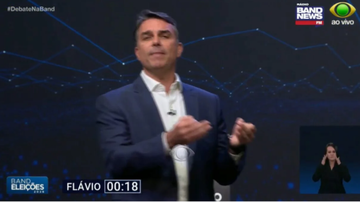 Flávio Bolsonaro durante o programa ao vivo, com fundo de tecnologia em tons escuros, microfone discreto no paletó e intérprete de Libras ao lado.